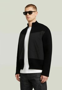 Giubbotto nero con zip, corpo in maglia testurizzata e accenti in pannelli lisci. Indossato sopra una t-shirt bianca, con jeans scuri e occhiali da sole.