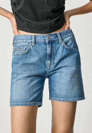 Personne portant un short en jean bleu clair et un crop top noir, les mains posées sur les hanches, sur un fond uni.