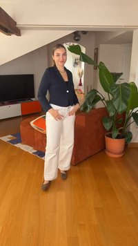 Νεαρή γυναίκα με μαύρο σακάκι και λευκό παντελόνι στέκεται χαμογελαστή σε ένα σαλόνι με ξύλινο πάτωμα, πορτοκαλί καναπέ, μεγάλο γλαστράκι και τηλεόραση.