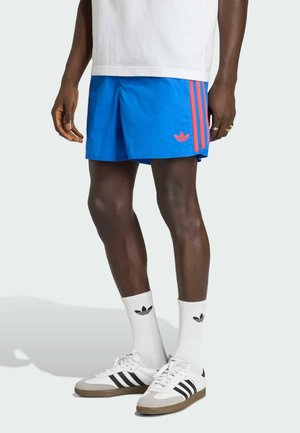 Blauwe sportbroek met roze strepen en logo, gecombineerd met witte sokken met zwart logo en witte schoenen met zwarte accenten.