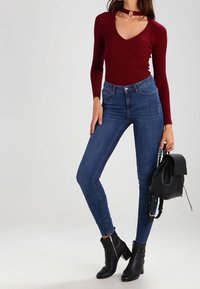 Burgundy pitkähihaisen ribbipaidan bodysuit, jossa on V-päänkaulus ja choker-tieto, yhdistettynä korkeavyötäröisiin sinisiin skinny-farkkuihin ja mustiin nilkkureihin.