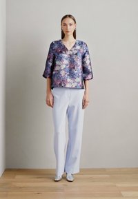 Haut à motif floral, en V, avec des manches trois-quarts dans des tons de violet et de bleu, associé à un pantalon large bleu clair et des chaussures argentées.