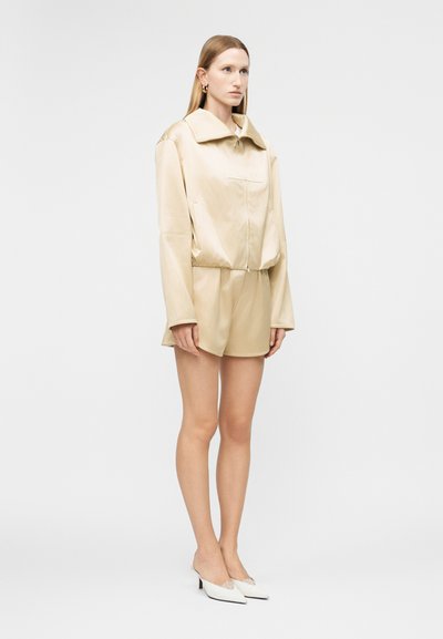 Γυναίκα που φοράει μπεζ υπερμεγέθη σακάκι και ταιριαστό σορτς με λευκές μυτερές γόβες, στέκεται μπροστά σε λευκό απλό φόντο.