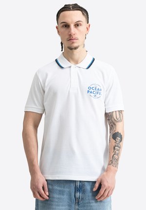 Muž s vrkočmi, oblečený v biele polo Ocean Pacific a modré džínsy, ukazuje detailné tetovania na ľavom predlaktí.