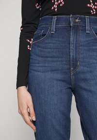 Jeans en denim bleu taille haute avec une texture lisse, cinq poches et des détails de couture orange. Portés avec un haut noir à manches longues.