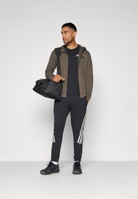 Ropa deportiva que incluye una sudadera con cremallera de color oliva oscuro, camiseta negra, pantalones de chándal negros con rayas blancas y zapatillas negras; llevando una bolsa de deporte negra.