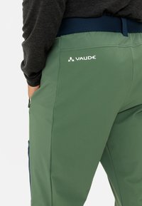 Groene broek met een gladde textuur, voorzien van een ritszak en een marineblauwe tailleband. Het "Vaude"-logo is zichtbaar op de achterkant.