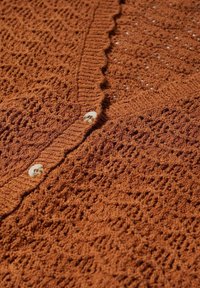 Pletený cardigan v rustikální oranžové barvě s texturovaným, otevřeným vzorem. Má vlnkovaný okraj a dva kulaté, krémové knoflíky.