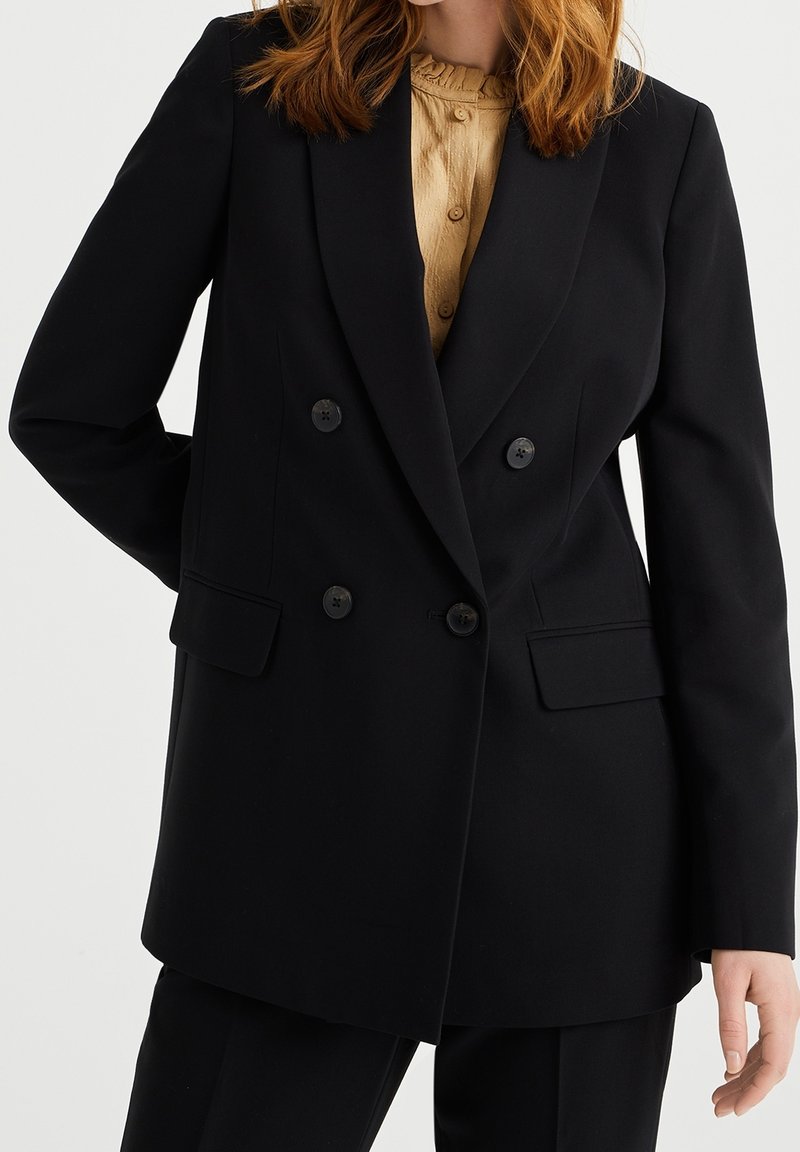 Blazer negro de doble botonadura con solapas muescas, dos bolsillos frontales y botones negros. La tela parece suave con un ajuste entallado.