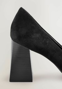 Zapato de tacón de gamuza negra con puntera afilada y un distintivo tacón angular de madera negra. Presenta una textura suave y un diseño minimalista.
