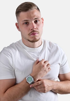 Silberner Armbanduhr mit grünem Zifferblatt, Zahlenmarkierungen und einem Edelstahlarmband, getragen am Handgelenk eines Mannes mit einem weißen T-Shirt.