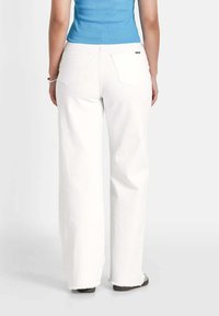Witte wijde jeans met een rafelige zoom, met twee achterzakken. Gekleed met een blauw aansluitend tanktop, gezien vanaf de achterkant.