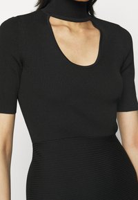 Haut noir côtelé avec un col roulé et un profond décolleté en U, présentant des manches courtes et une silhouette ajustée. La texture est lisse avec des lignes verticales.