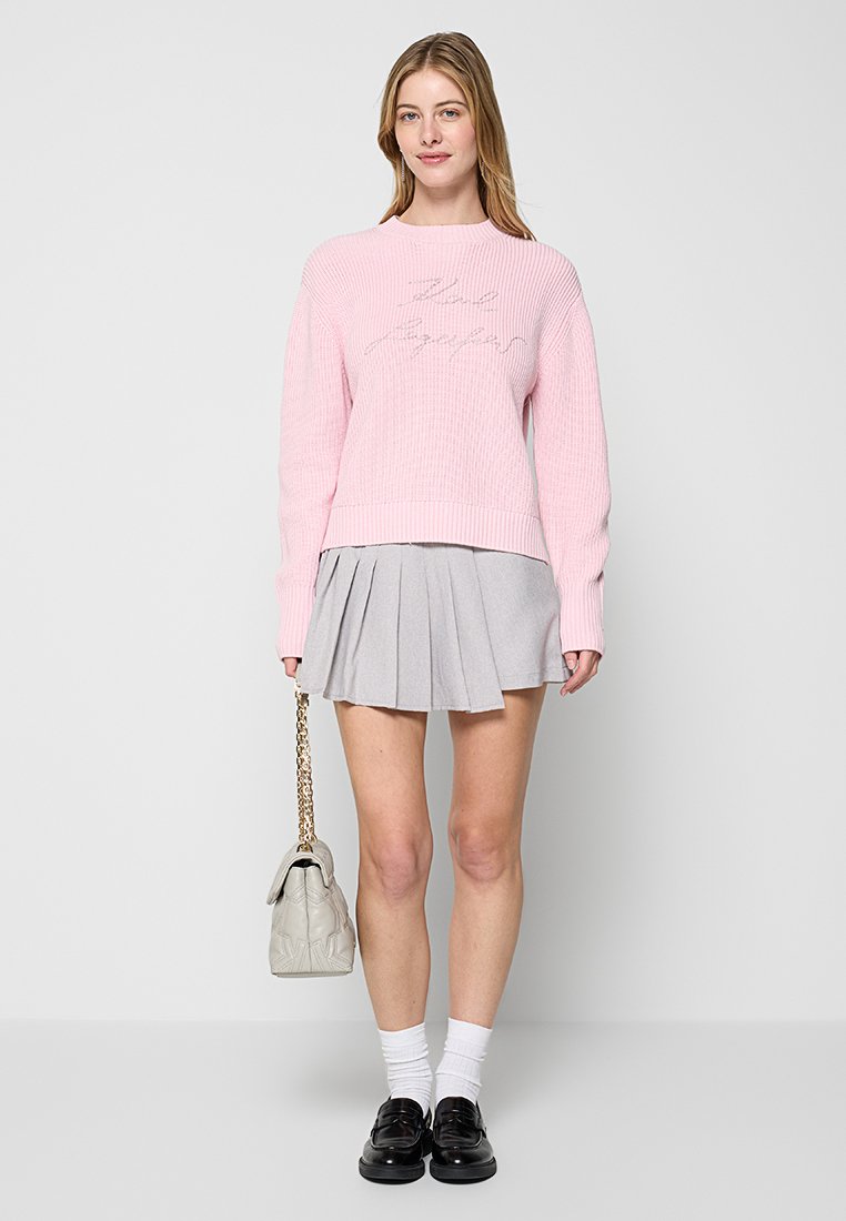 Pull en tricot côtelé rose clair avec texte imprimé, associé à une jupe plissée grise. Les accessoires incluent un sac à main gris et des chaussures noires.