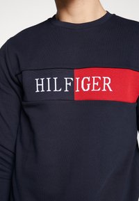 Personne portant un sweat-shirt bleu marine avec un panneau horizontal rouge et bleu affichant le texte blanc "HILFIGER" sur la poitrine.