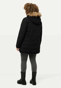 Svart puffjacka med huva i syntetpäls, med mellanhöjdsklipp och sidofickor, kombinerad med gråjeans och svarta stövlar.