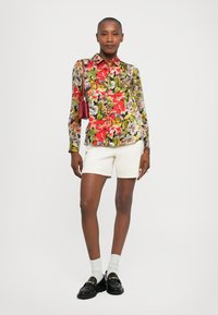 Cămașă florală în roșu, verde și galben, asortată cu pantaloni scurți albi. La acest outfit se adaugă pantofi loafers negri, ciorapi albi și o geantă mică roșie.