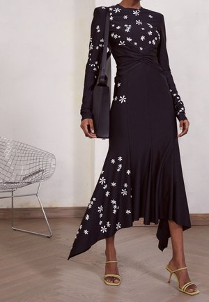 Schwarzes langärmeliges Kleid mit weißer Blumenstickerei, tailliertem Oberteil und asymmetrischem Saum. Kombiniert mit grünen Riemchenabsätzen.