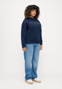 Felpa blu marina con logo frontale, cappuccio regolabile con cordini e polsini a costine, abbinata a jeans azzurri larghi e scarpe da tennis bianche.