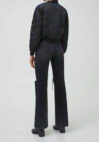 Veste bomber noire avec des poignets côtelés, assortie à un jean noir taille haute et jambes larges, affichant un ourlet brut et un détail zippé sur le côté.