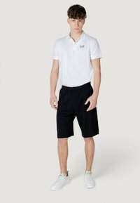 Polo bianco con maniche corte e logo, abbinato a pantaloni neri e sneakers bianche. Design casual e atletico.