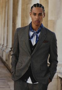 Gray herringbone suit with a black vest and white shirt, accented by a blue patterned scarf. Model has braided hair and slight facial features.