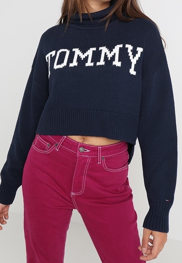 Marineblauer gestrickter, verkürzter Pullover mit weißem Schriftzug "TOMMY"; kombiniert mit hoch taillierten, rosa Cordhosen mit Kontrastnähten.