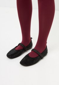 Zapatos planos negros con puntera redonda, adornados con brillantes strass y una correa negra texturizada, usados sobre medias burdeos.