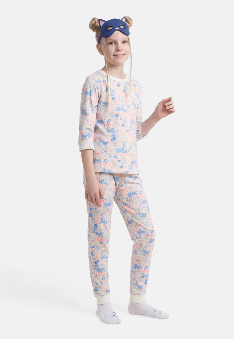 Jeune fille portant un pyjama pastel à motifs de cerfs et de fleurs, un masque bleu en forme de chat et des chaussettes blanches avec de petits visages d'animaux.