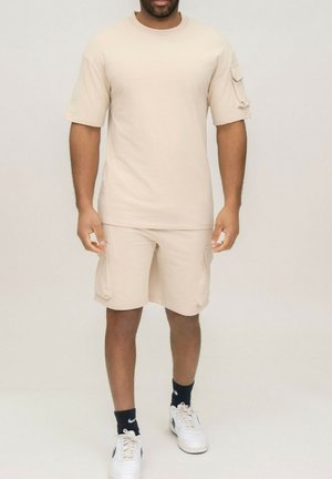 Homme portant une chemise beige à manches courtes et un short cargo assorti, avec des baskets blanches et des chaussettes noires Nike, debout devant un fond uni.