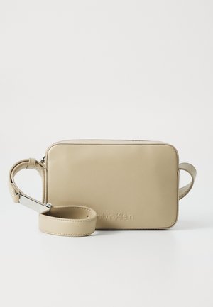 Béžová obdélníková kabelka Calvin Klein přes tělo s nastavitelným popruhem položená vpředu, jednoduchý design, hladká textura a viditelné značení značky.