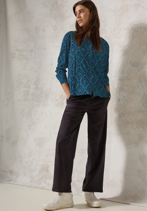Camicia a pattern blu con design a diamante, abbinata a pantaloni neri a gamba larga e sneaker sportive bianche, in piedi davanti a uno sfondo neutro.