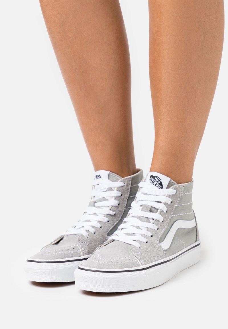 Personne portant des baskets montantes Vans grises avec des lacets et des semelles blanches, debout sur une surface blanche unie.