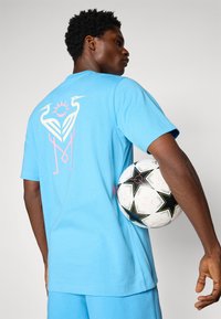 Light blue cotton t-shirt featuring a large pink and white graphic of two birds and a sun on the back; model holds a black and white soccer ball.