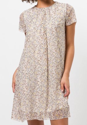 Kobieta ubrana w sukienkę z krótkim rękawem, sięgającą do kolan, z lekkiego materiału o drobnym, brązowo-żółto-pomarańczowym wzorze kwiatowym na białym tle.