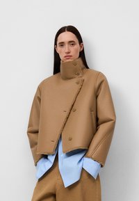 Braune, taillierte Jacke mit hohem Kragen, großen Knöpfen und rohen Kanten. Wird über einem hellblauen Hemd getragen. Glatte Textur, passgenauer Schnitt.