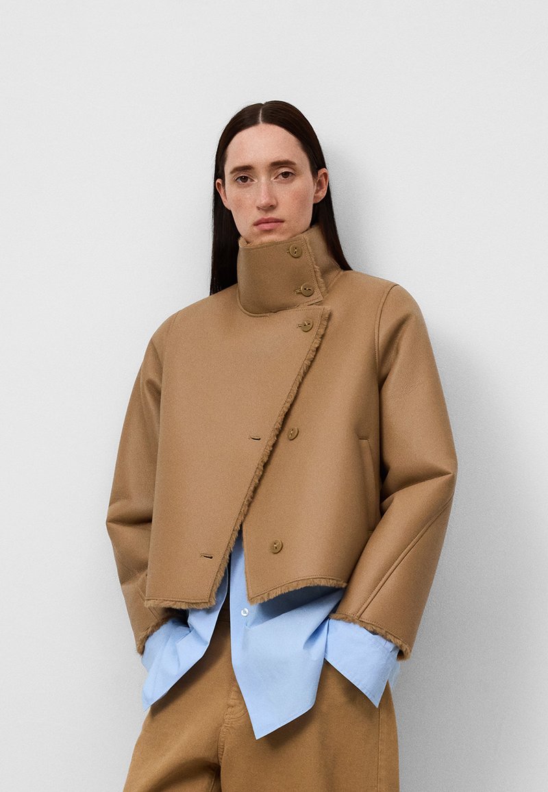 Braune, taillierte Jacke mit hohem Kragen, großen Knöpfen und rohen Kanten. Wird über einem hellblauen Hemd getragen. Glatte Textur, passgenauer Schnitt.