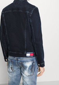 Tumman sininen farkkutakki klassisella kauluksella, ommellulla yksityiskohdalla ja Tommy Jeans -logopatchilla. Yhdistetty vaaleasuhtisiin kulutettuihin farkkuihin.