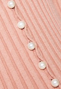 Jasnoróżona prążkowana dzianina z perełkowymi zdobieniami wzdłuż falistego szwu ułożonego pionowo, tworząca teksturowany element.