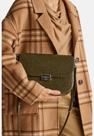 Bolso de mano verde con textura de cocodrilo, cierre de solapa y herrajes plateados, sostenido contra un abrigo de cuadros marrón con una parte superior color beige.