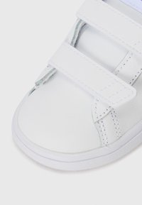 Baskets blanches avec une empeigne en cuir lisse, deux sangles Velcro, un bout arrondi et une semelle en caoutchouc texturée pour une meilleure adhérence. Détails de couture visibles.