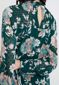 Zielona sukienka w kwiaty z miękkiej, teksturowanej tkaniny. Posiada otwarcie w kształcie łezki z tyłu oraz długie, zwiewne rękawy ozdobione kropeczkami.