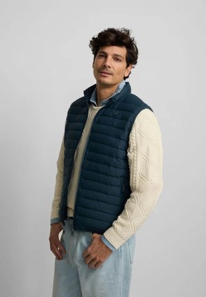 Hombre con vaqueros azul claro, suéter de punto grueso color crema y chaleco sin mangas acolchado azul marino, de pie frente a un fondo gris liso.