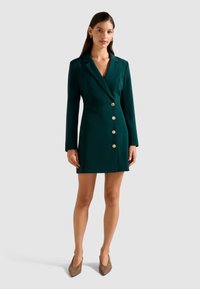 Donna che indossa un blazer dress verde scuro aderente con bottoni dorati, maniche lunghe e décolleté beige a punta, in piedi contro uno sfondo neutro.