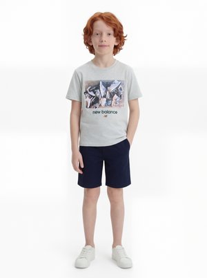 Rapaz ruivo está de pé, vestindo uma t-shirt cinza clara da New Balance com gráfico de ténis, calções azul escuro e ténis brancos, sobre fundo branco.