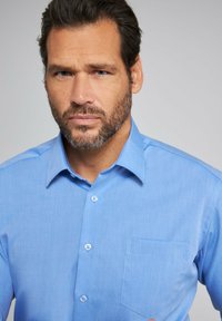 Camisa de botones de color azul claro, hecha de una tela suave. Presenta un collar, un bolsillo en el pecho y cinco botones a juego en la tapeta.