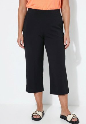 Femme portant un pantalon noir court à jambes larges, un haut orange et des sandales plates tressées, debout sur un fond blanc.