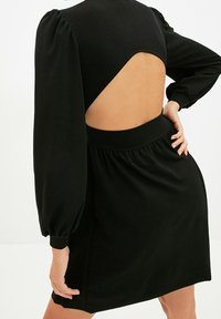 Czarna sukienka z otwartym tyłem, długimi bufkami, dopasowanym pasem i gładką teksturą materiału. Podkreślona marszczonym pasem.