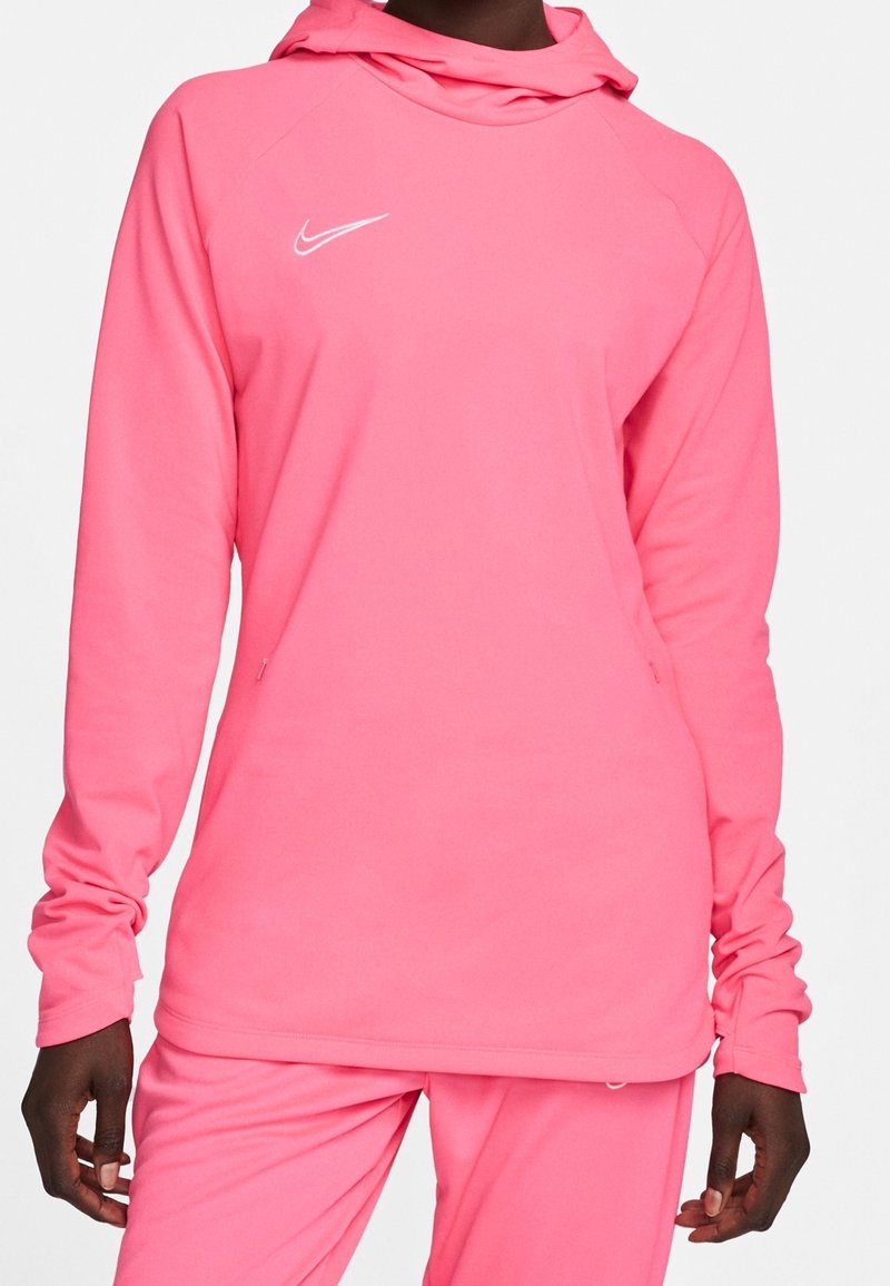 Personne portant un haut à capuche à manches longues Nike rose vif et un pantalon assorti, vue de face avec le logo Nike blanc sur la poitrine.