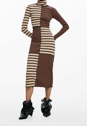 Femme portant une robe midi à manches longues avec des empiècements à rayures verticales en marron, beige et noir, associée à des bottines noires décontractées.